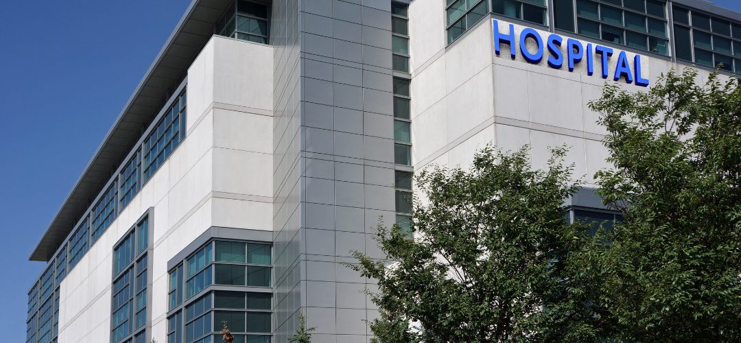 Exterior view of a modern hospital building with large glass windows and the word "HOSPITAL" displayed prominently in blue letters.