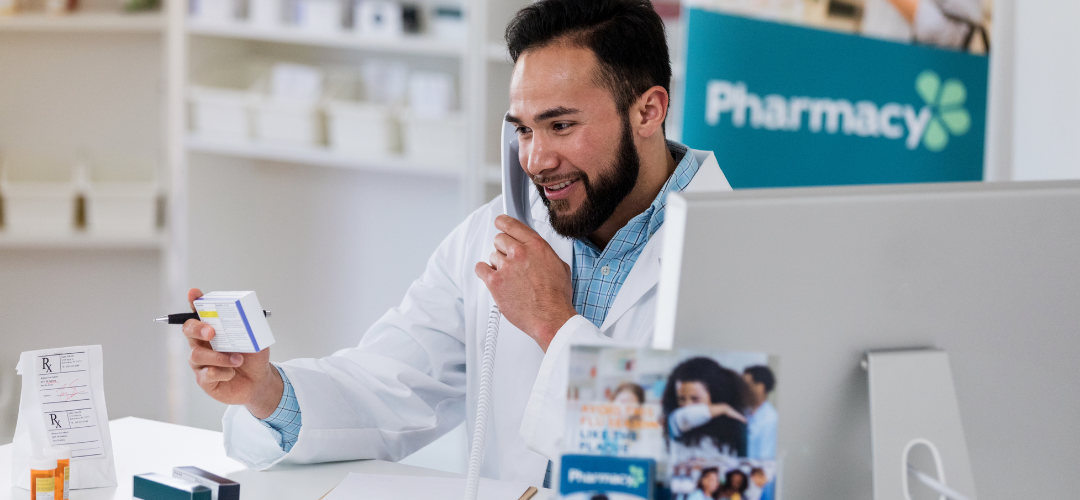 Pharmacist holding a medication box while speaking on the phone, with pharmacy-related items and signage in the background.